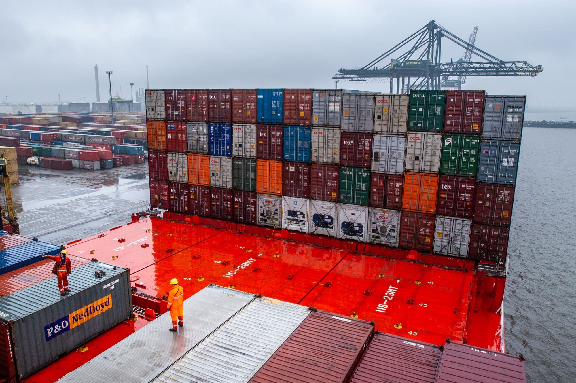 Trabalhadores portuários com equipamento de proteção sobre o convés de um navio porta-contêineres, ao lado de pilhas de contêineres e guindastes